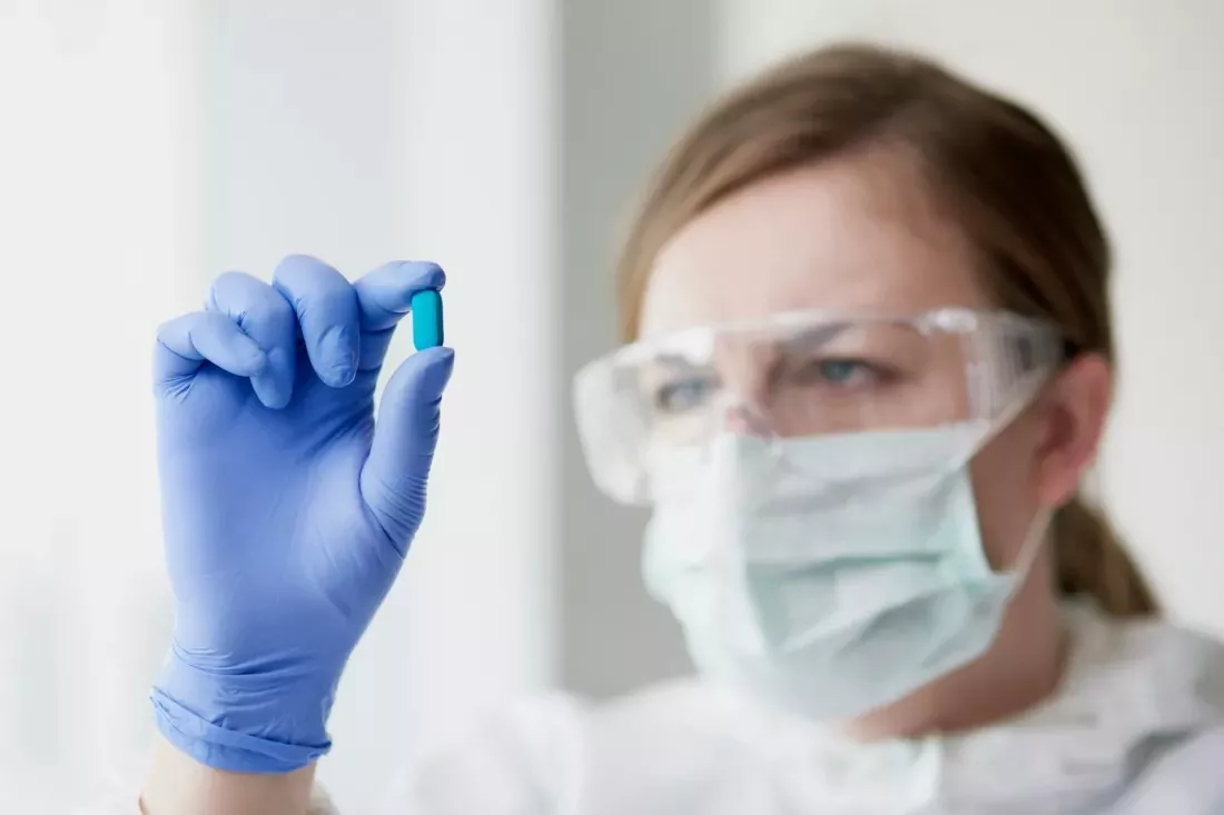 A lab technician in safety goggles and a mask looks at a blue capsule.