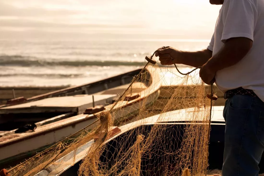 Fisherman untangling a fishing net next to boats on a beach at sunset