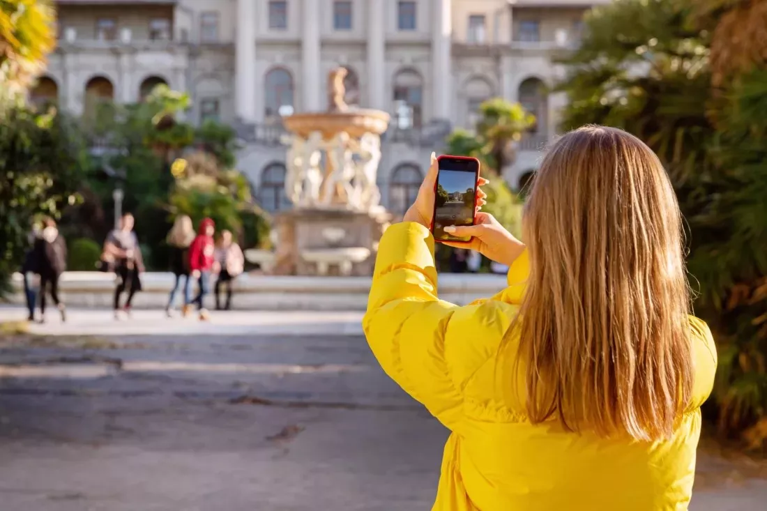 Young woman in a yellow jacket photographs a city square with her phone